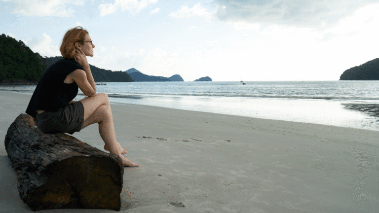 Nômade digital pensativo olhando o pôr do sol na praia, refletindo sobre os desafios da saúde mental ao viajar sozinho.